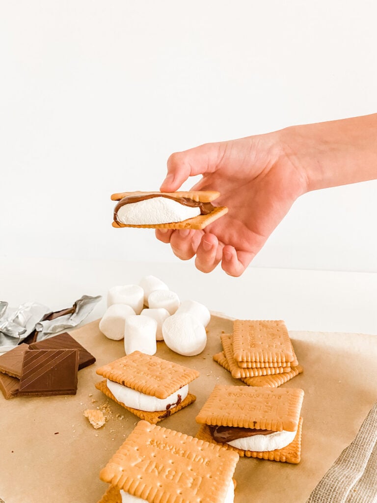 A hand holding an Everyday Oven S'mores against a white background and over all the ingredients