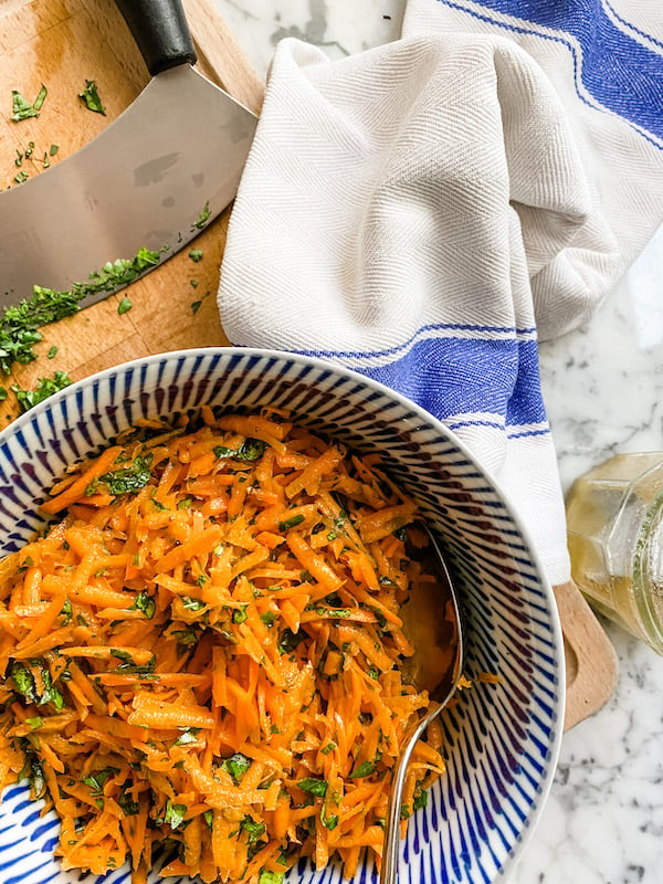 Grated carrot salad in a blue bowl with chopped herbs, a knife and a white tea towel