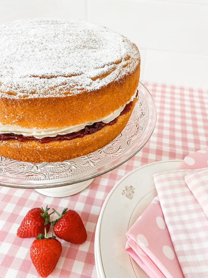 A Victoria Sponge Cake on a glass cake stand, standing on a table with a pink gingham tablecloth