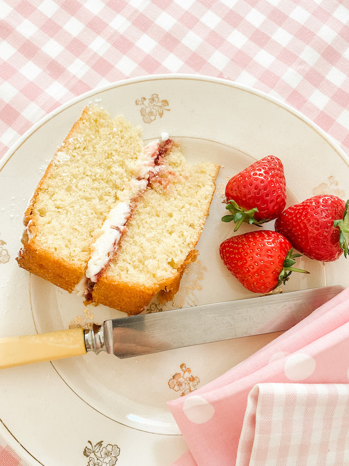 Slice of Victoria Sponge Cake and 3 strawberries on a plate with a knife and pink gingham napkins