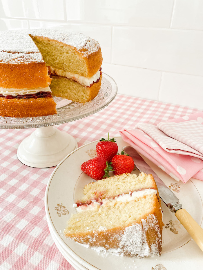 A Victoria Sponge Cake on a glass cake stand, standing on a table with a pink gingham tablecloth