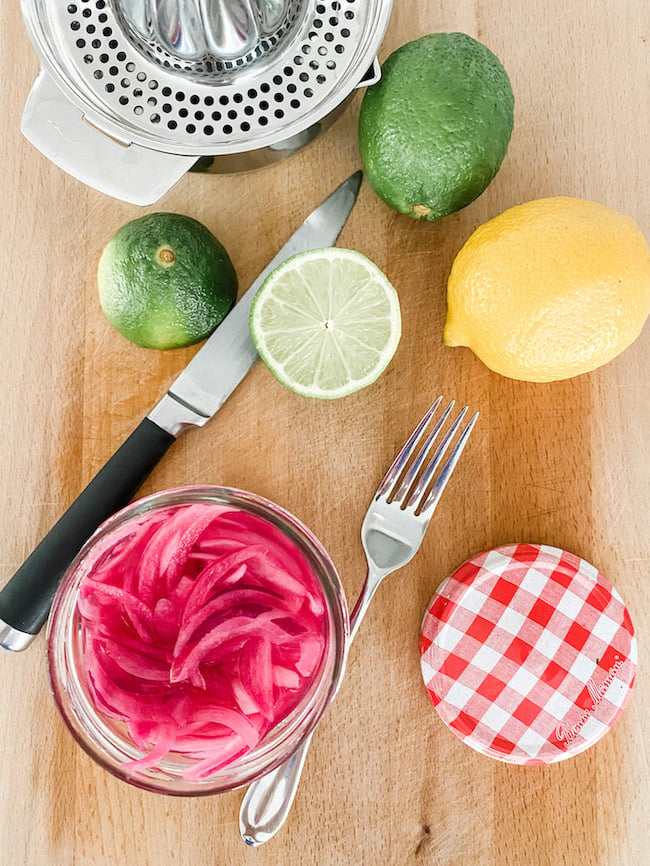 Pickled Red Onions in a jar on a chopping board with limes and a lemon